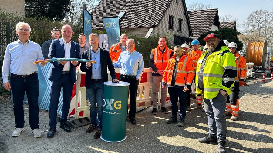 Gruppe von Personen und Bauarbeitenden steht bei einem symbolischen Baustart neben einer Absperrung und einer OXG‑Säule im Außenbereich.