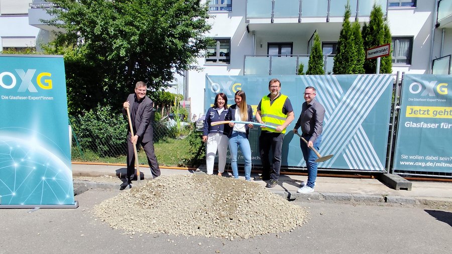 Eröffnung eines Glasfaserprojekts in Augsburg mit mehreren Personen, die Schaufeln halten und sich neben einem großen Erdhaufen und einem Werbebanner aufstellen
