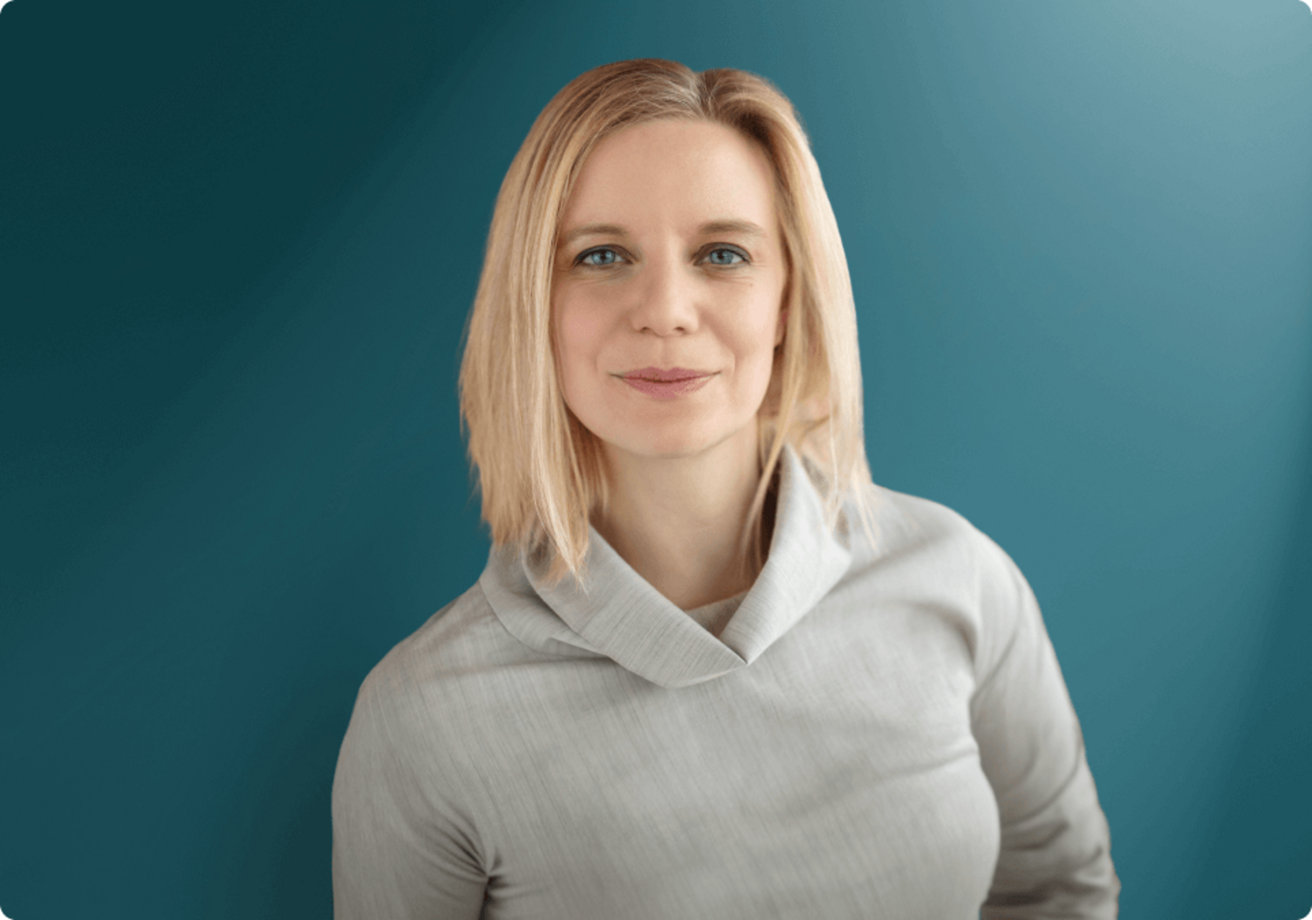 Porträt einer lächelnden Frau mit blonden Haaren, die ein graues Oberteil trägt, vor einem dunkelblauen Hintergrund