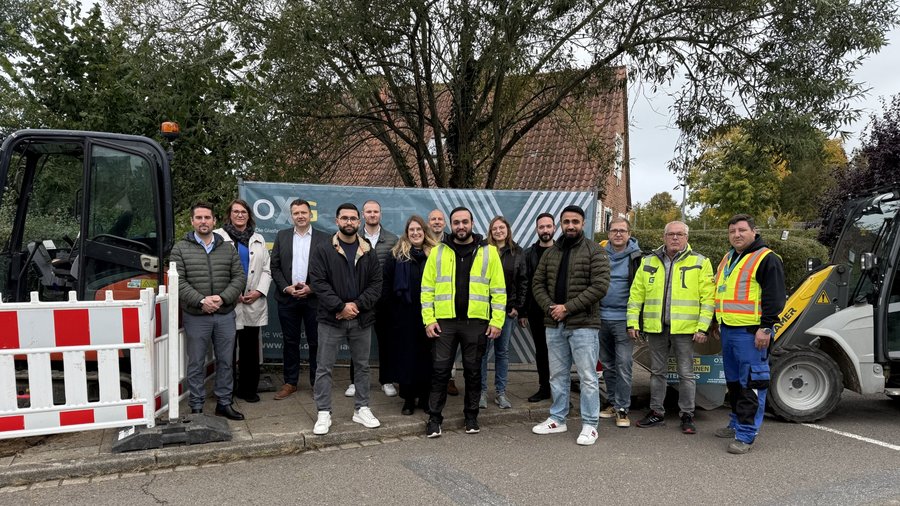 Gruppe von Fachleuten in Arbeitskleidung stehen zusammen vor einem Bagger und einem Banner, umgeben von Bäumen und einem Wohngebäude in Kaltenkirchen.