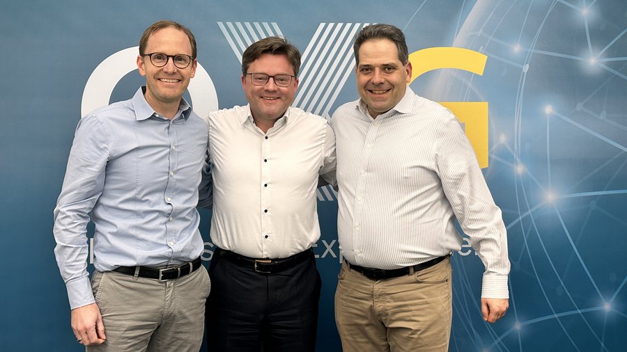 Drei Männer stehen lächelnd nebeneinander vor einem blauen Hintergrund mit grafischen Elementen und dem Text "OXG".