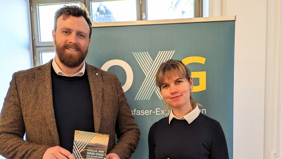Zwei Personen, ein Mann und eine Frau, stehen vor einem Banner mit dem Logo von "OXG" und halten Informationen über Glasfaser-Expansion in den Händen.