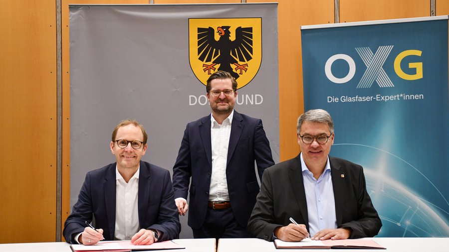 Drei Männer sitzen an einem Tisch und unterzeichnen Dokumente, während im Hintergrund das Wappen von Dortmund und ein Banner mit dem Logo von OXG zu sehen ist
