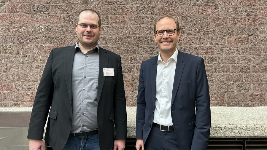 Zwei Männer in formeller Kleidung stehen nebeneinander vor einer roten Backsteinwand.