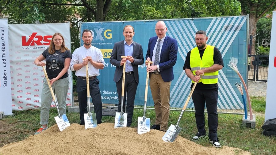 Gruppenfoto von fünf Personen, die an einem Spatenstich für ein Bauprojekt in Berlin Steglitz-Zehlendorf teilnehmen, umgeben von Sand und Werbebannern der beteiligten Firmen