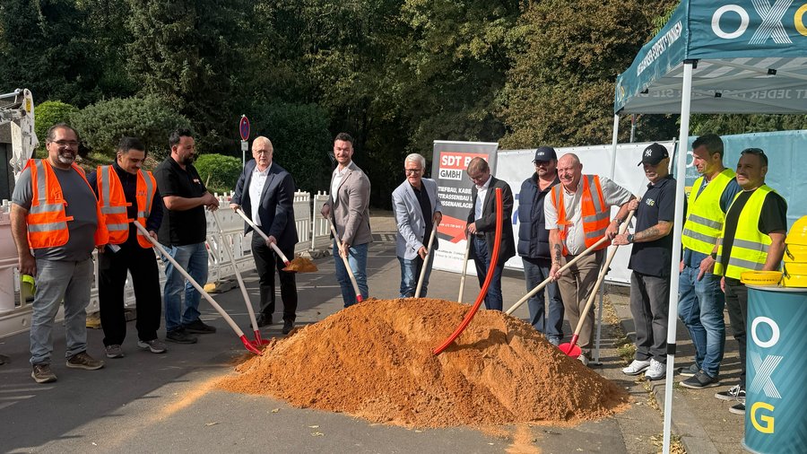 Mehrere Personen stehen mit Spaten um einen Sandhaufen auf einer abgesperrten Straße und setzen gemeinsam einen symbolischen ersten Spatenstich für ein Infrastrukturprojekt um.