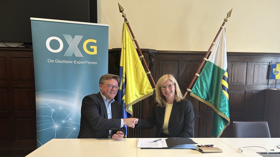 Zwei Personen schütteln sich die Hände vor einem Banner der OXG, während im Hintergrund Fahnen und ein Tisch mit Dokumenten sichtbar sind.