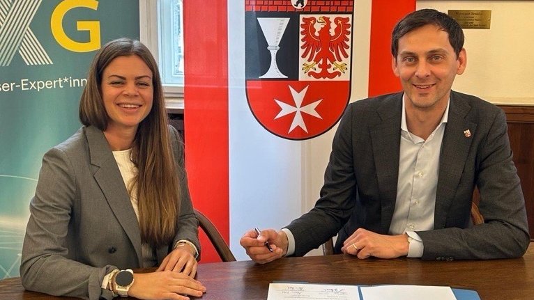 Zwei Personen sitzen an einem Tisch, lächeln in die Kamera und sind vor einer Flagge sowie einem Wappen positioniert, während sie ein Dokument zur Unterzeichnung bereit haben
