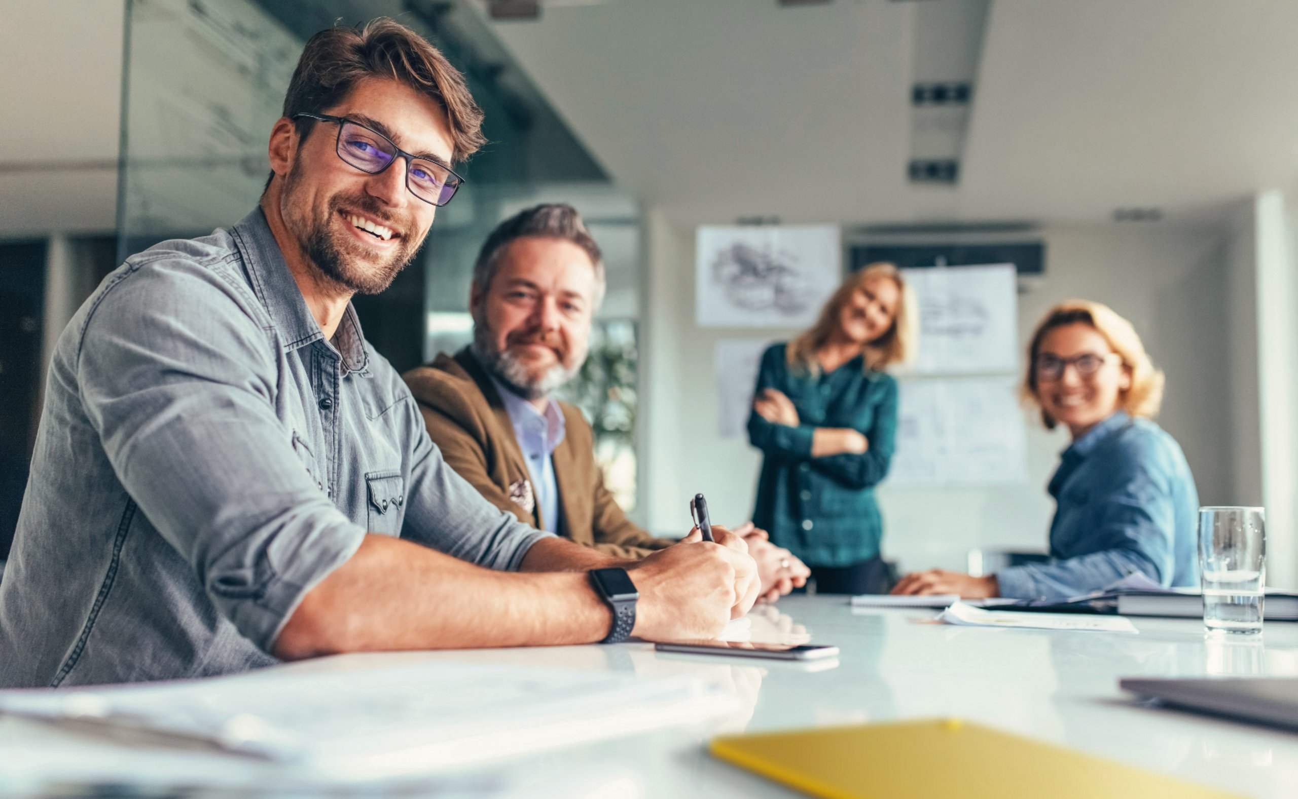 Teamarbeit in einem modernen Büro: Eine Gruppe von vier Personen, bestehend aus zwei Männern und zwei Frauen, sitzt an einem Tisch und diskutiert, während sie in die Kamera lächeln.