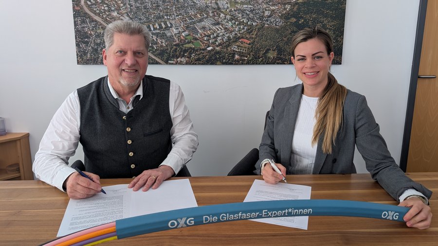 Ein Mann und eine Frau sitzen an einem Tisch in einem Büro und unterschreiben Dokumente; davor liegt ein OXG‑Glasfaserkabel, im Hintergrund ein Wandbild mit einer Luftaufnahme der Stadt Neuried.