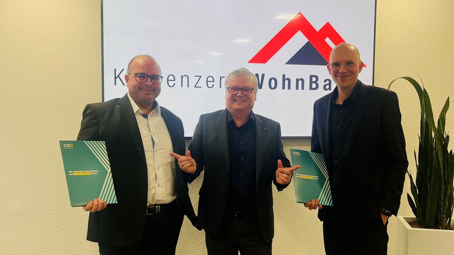 Drei Männer in Anzügen halten Ordner in der Hand und posieren vor einem Unternehmenslogo mit dem Schriftzug "Konzernz WohnBau"