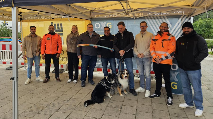 Gruppenfoto von Mitarbeitern und Hunden unter einem Pavillon, umgeben von Materialien zur Datenübertragung, an einem öffentlichen Ort
