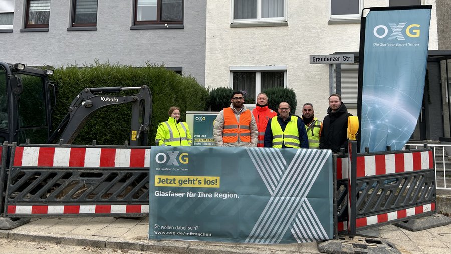 Gruppe von Personen in Arbeitskleidung mit einem Bagger hinter einer Baustellenabsperrung, während sie für ein Glasfaserprojekt in der Graudenzer Straße in Bremerhaven posieren