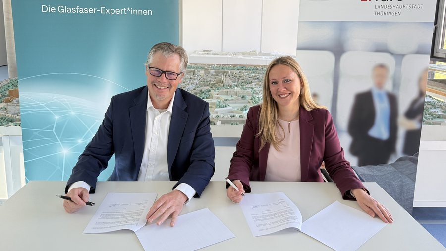 Zwei Personen sitzen an einem Tisch und unterzeichnen Verträge. Im Hintergrund ist eine Präsentation mit einem Modell der Stadt Erfurt und einem Banner mit dem Schriftzug „Die Glasfaser-Expert*innen“ zu sehen.