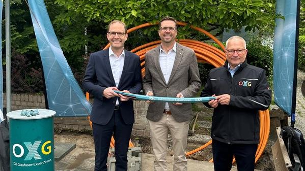 Drei Personen stehen in der Stadt Köln im Freien und halten gemeinsam ein Stück Schlauch, während im Hintergrund orangefarbene Kabelrollen und ein Stand mit dem Logo von OXG sichtbar sind