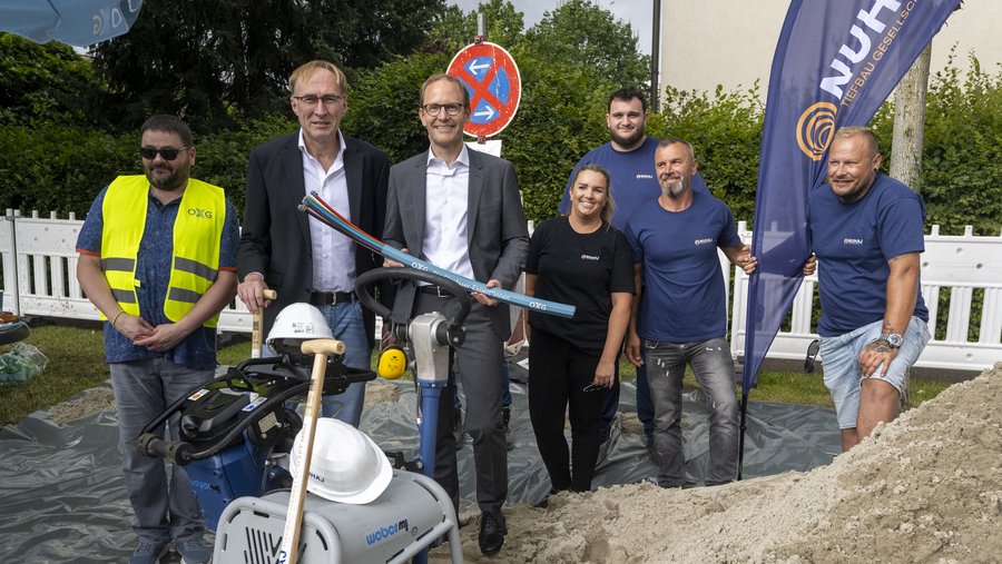 Gruppenfoto von fünf Personen in Arbeitskleidung und Schutzwesten, die sich um eine Baustelle in Freiburg mit einem Baugerät und Materialien versammeln, während ein Baustellenbanner im Hintergrund zu sehen ist