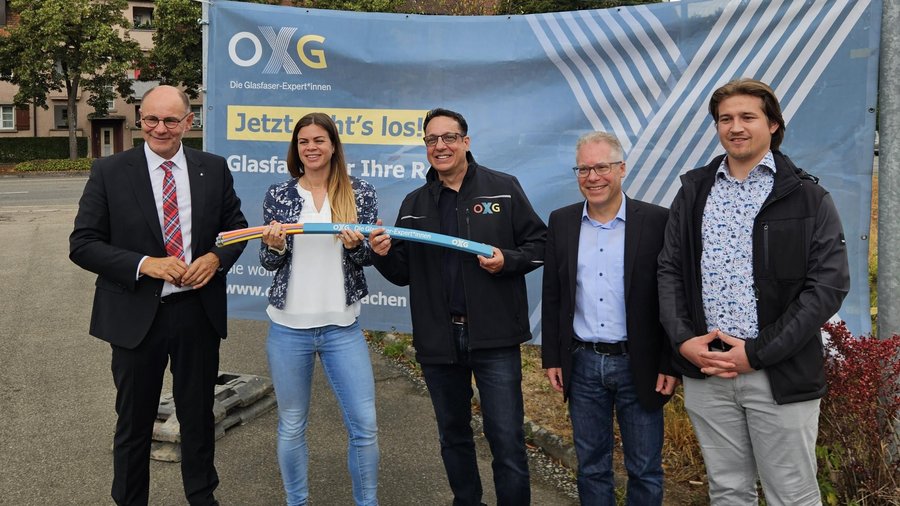 Fünf Personen stehen in Singen vor einem Werbebanner von OXG und halten ein Stück Glasfaserleitungen in den Händen.