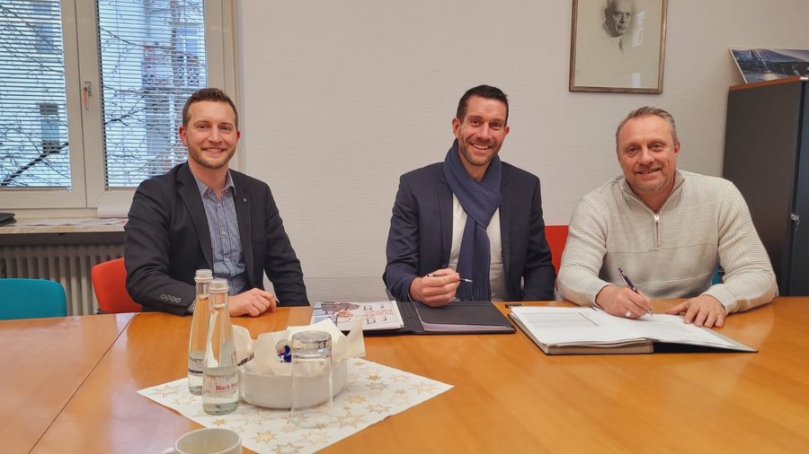 Drei Männer sitzen an einem Tisch in einem Büro, lächeln und sind mit Schreibmaterialien beschäftigt, während Wasserflaschen und eine Unterlage auf dem Tisch stehen