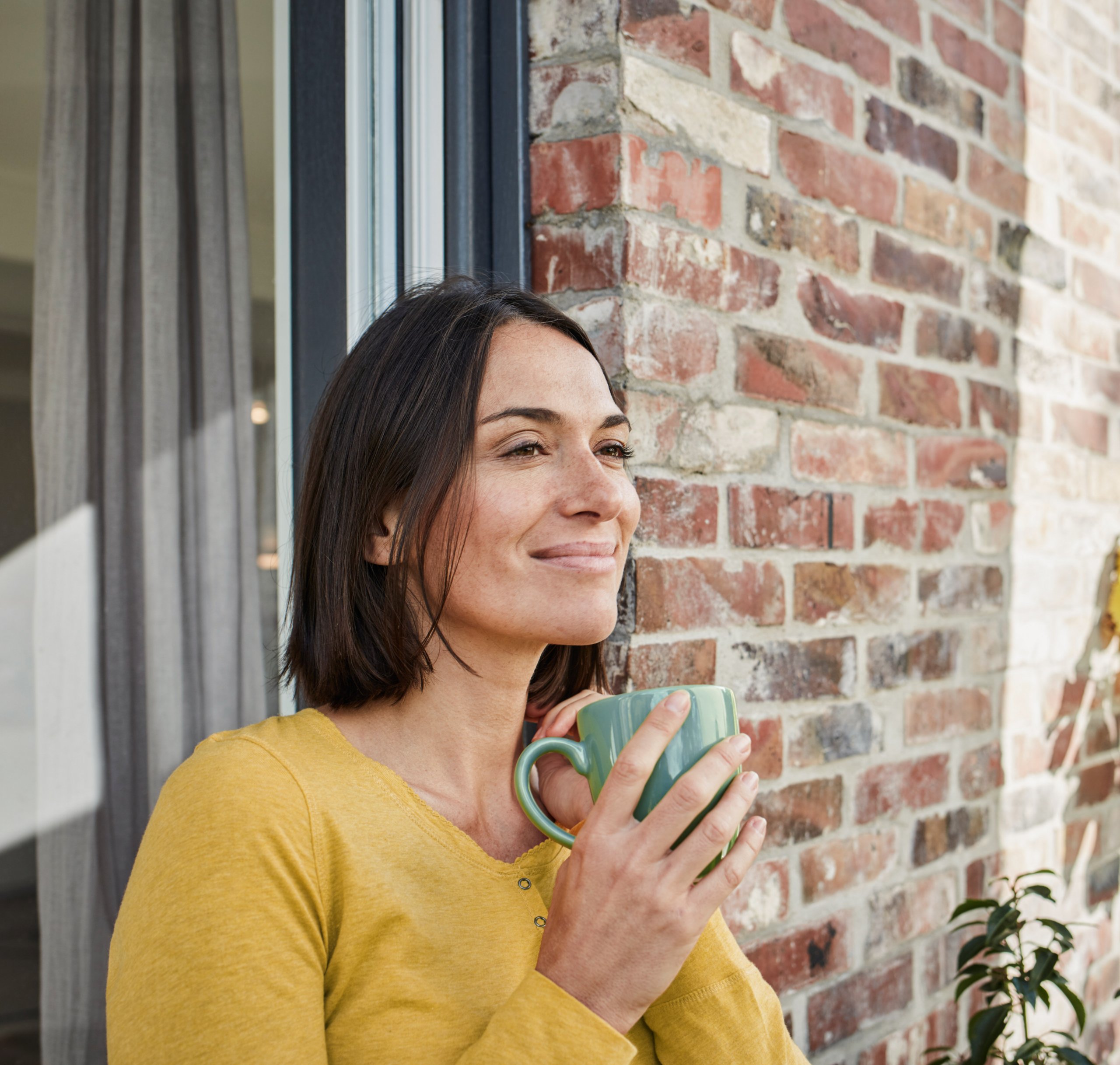 Eine Frau in einem gelben Sweatshirt hält eine grüne Tasse und steht an einem Fenster, während sie nachdenklich lächelt