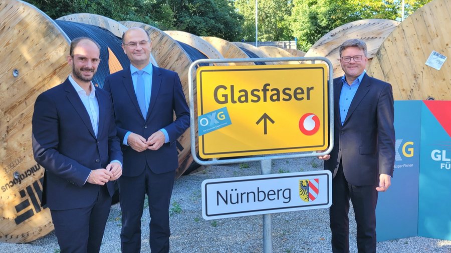 Drei Personen stehen vor einem Straßenschild mit der Aufschrift „Glasfaser“ sowie dem Logo von Vodafone, im Hintergrund sind große Kabeltrommeln zu sehen.