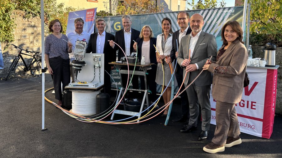 Ein Gruppenfoto von neun Personen unter einem OXG‑Infostand im Freien. Sie stehen vor technischen Komponenten und Kabeln für den Glasfaser‑Ausbau und halten farbige Leitungen in der Hand. Im Hintergrund sind ein Banner mit Firmenlogos, ein Wohnhaus, Herbstbäume sowie Fahrräder zu sehen, rechts ein Stehtisch mit Kaffeekannen und Gläsern.