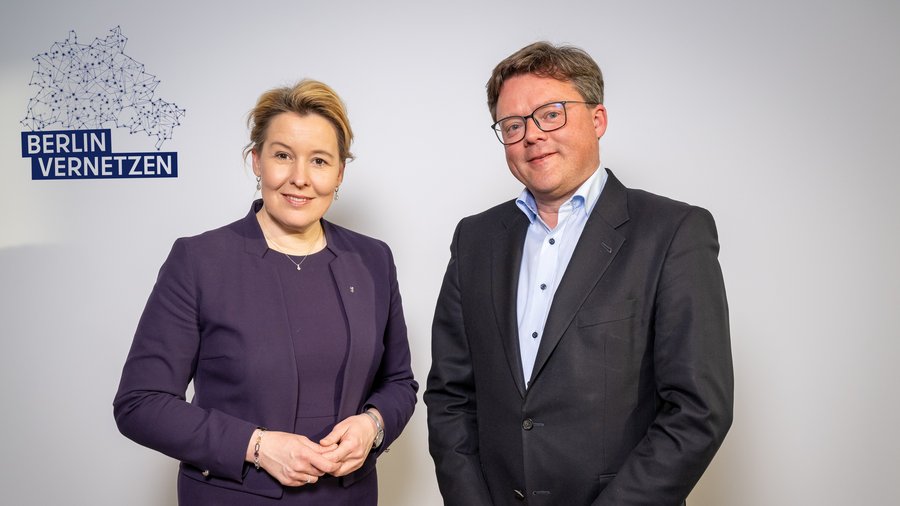 Zwei Personen, eine Frau in einem dunklen Blazer und ein Mann in einem Anzug, posieren lächelnd vor einem weißen Hintergrund mit dem Schriftzug "BERLIN VERNETZEN" und einer grafischen Darstellung von Netzwerken