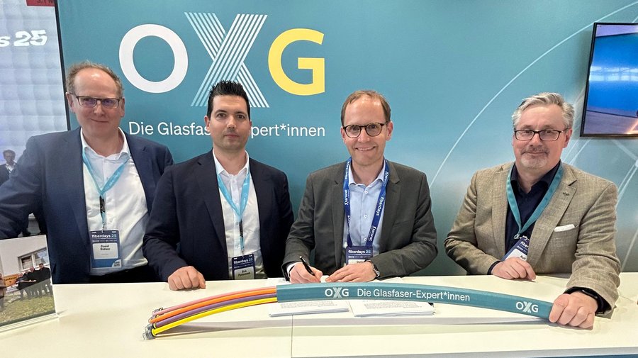 Vier Männer stehen an einem Tisch, auf dem eine bunte Glasfaserleitung und ein Schild mit der Aufschrift "OXG - Die Glasfaser-Expert*innen" zu sehen sind, während im Hintergrund eine Werbefläche zu erkennen ist