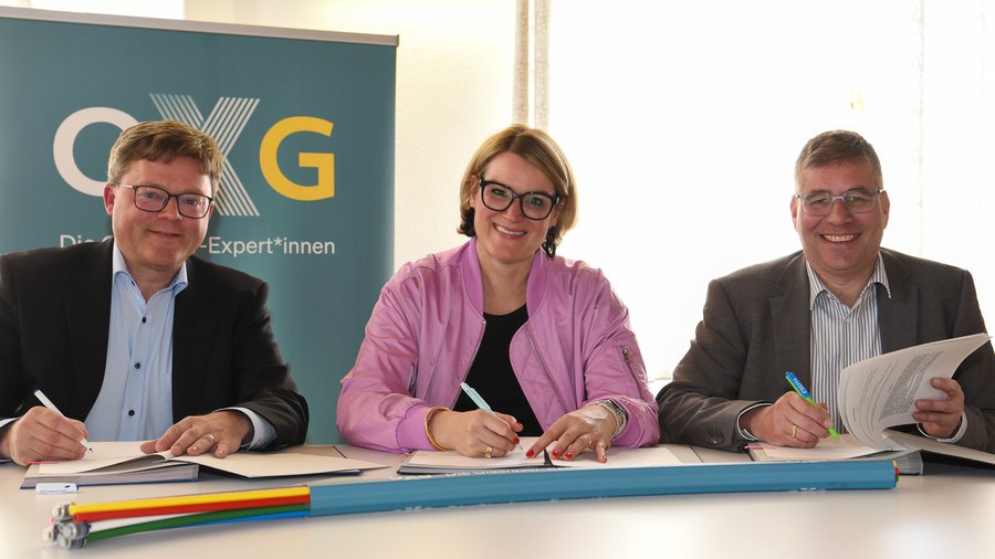 Drei Personen sitzen an einem Tisch, unterzeichnen Dokumente zum Glasfaserausbau in der Region Augsburg und lächeln, während im Hintergrund ein Banner mit dem Schriftzug "OXG - Die digitalen Expert*innen" zu sehen ist.
