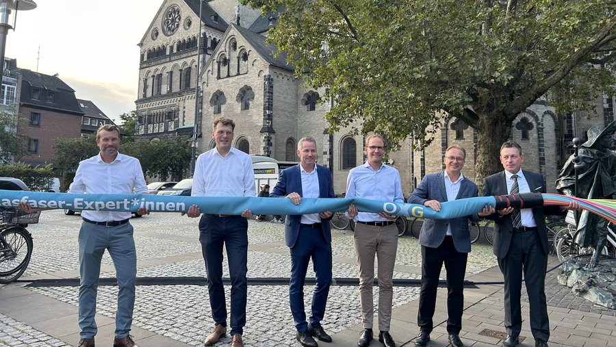 Sechs Personen, die ein großes Kabel mit der Aufschrift "Glasfaser-Expert*innen" halten, stehen vor einer historischen Gebäudeansicht in einem Stadtzentrum in Neuss