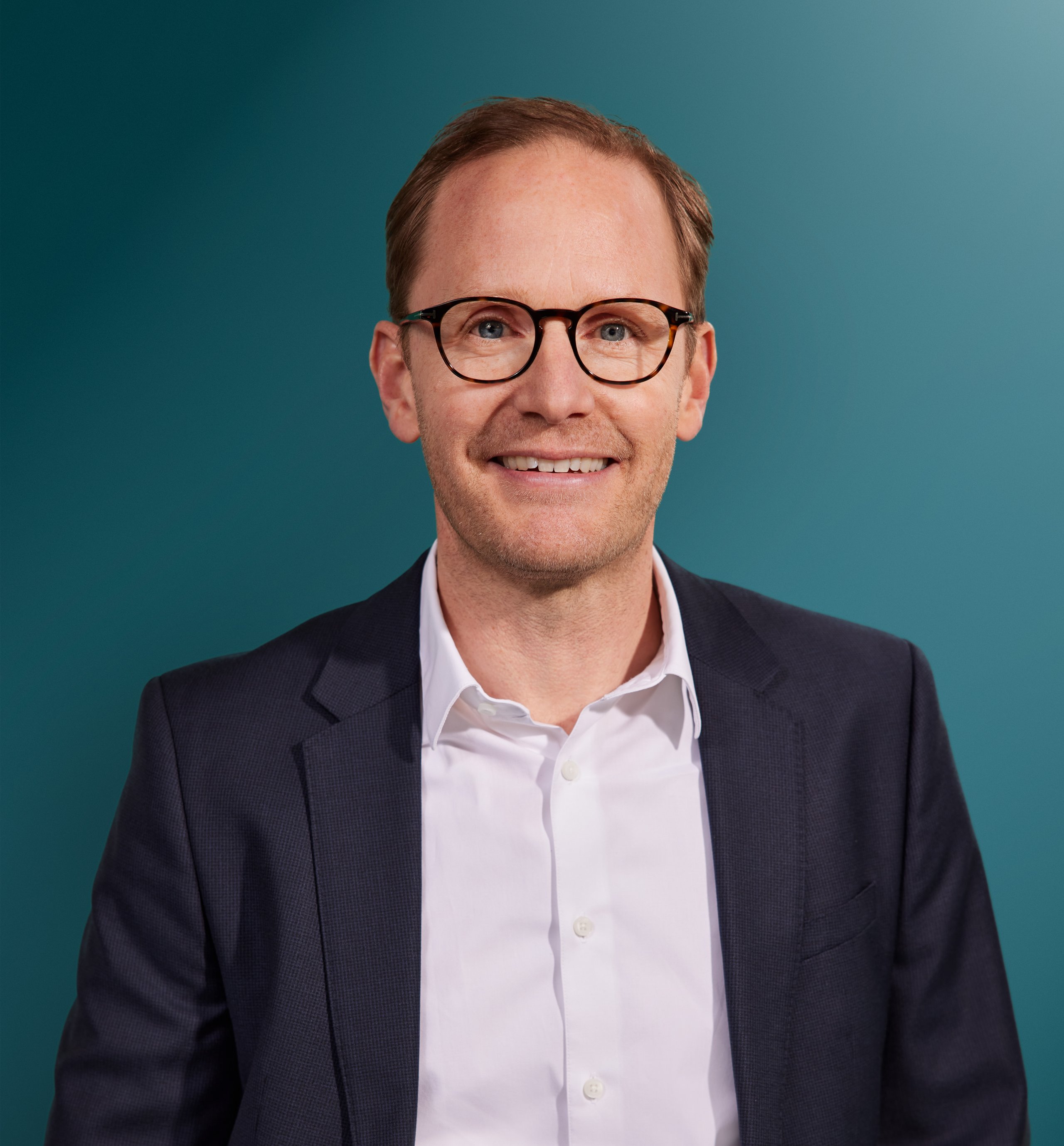 Porträt eines lächelnden Mannes in Anzugjacke und Hemd vor einem einfarbigen Hintergrund