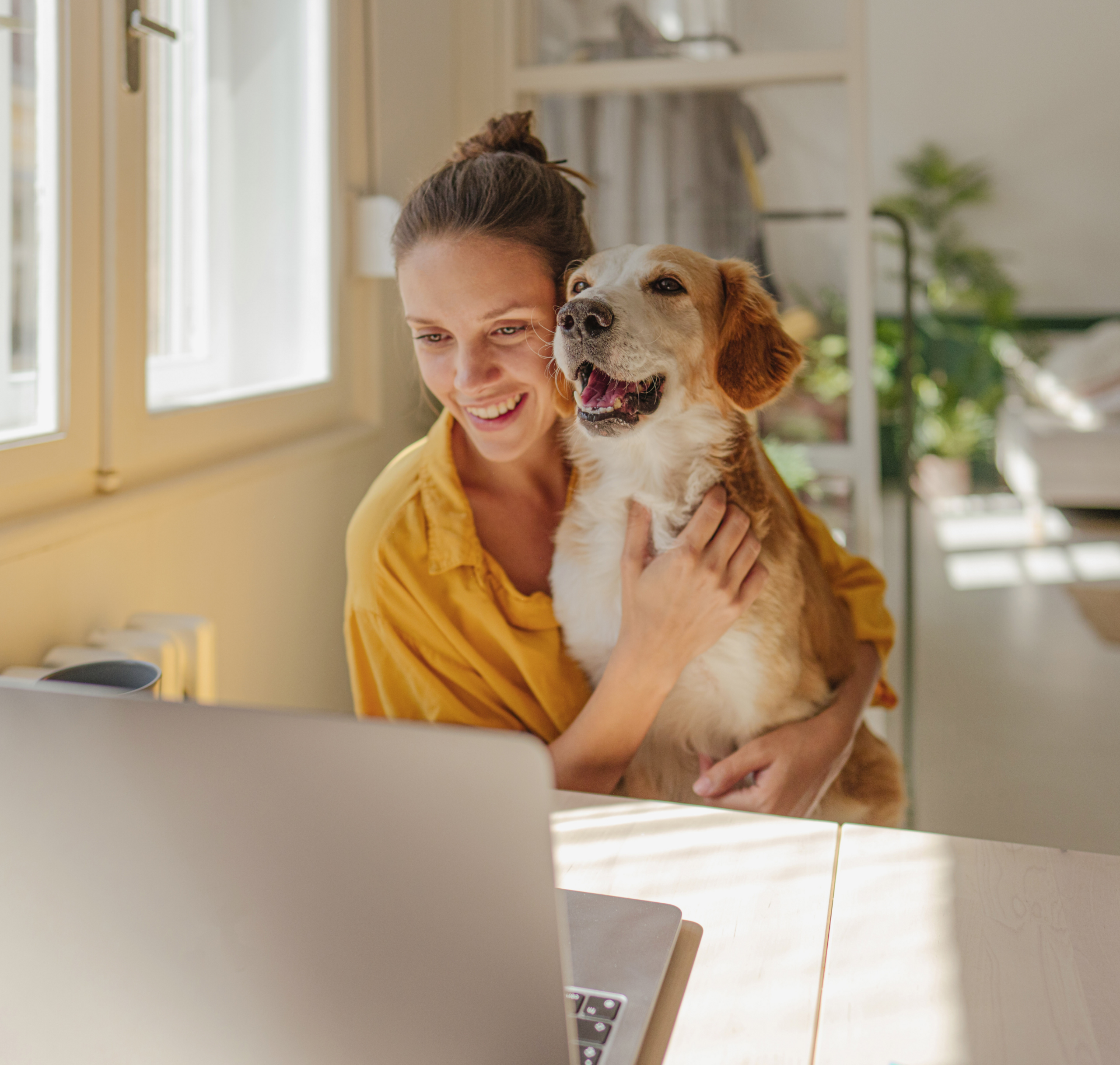 Eine Frau mit einem Hund sitzt an einem Tisch und schaut lächelnd auf einen Laptop