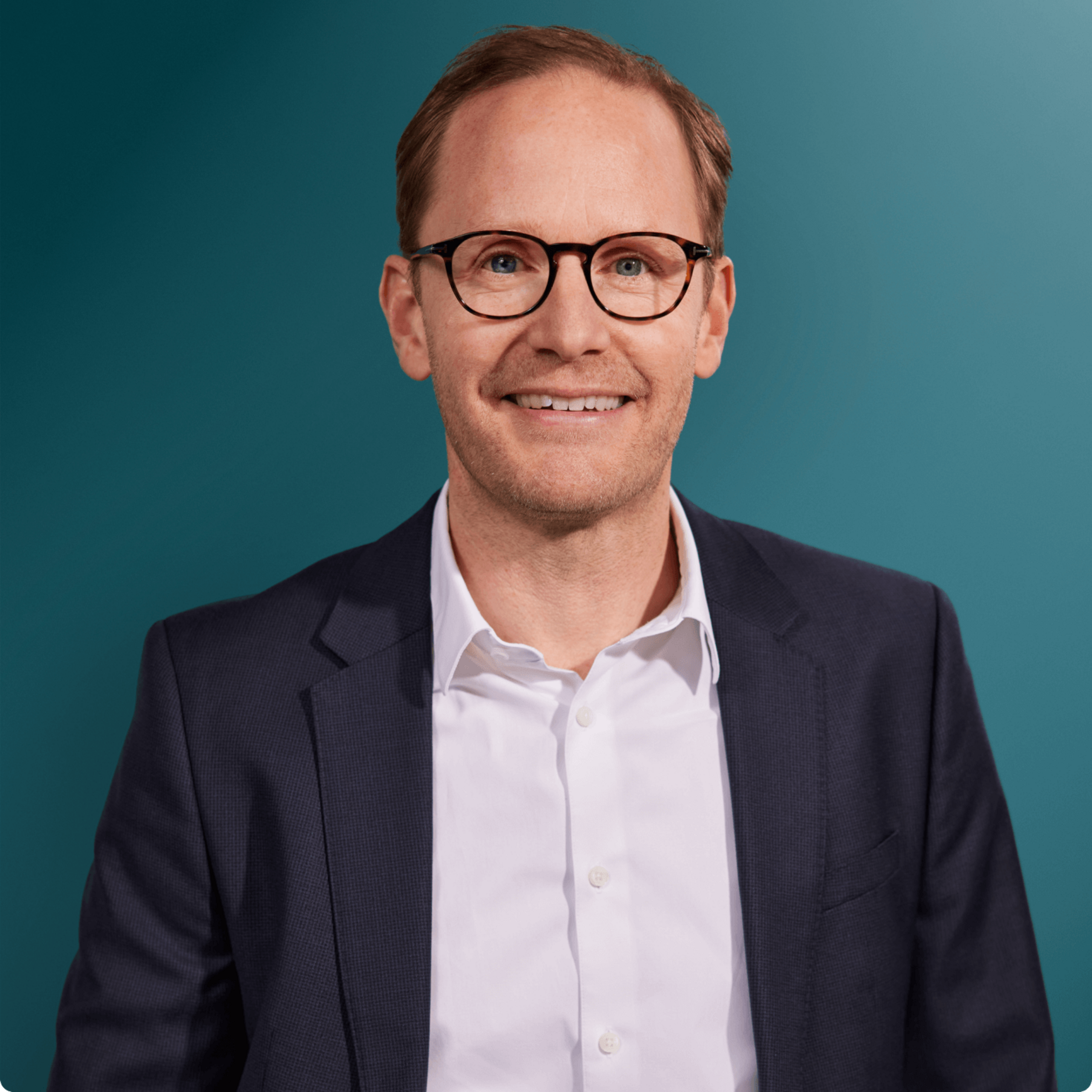 Portraitaufnahme eines Mannes mit Brille, der in einem dunklen Anzug und einem weißen Hemd vor einem blauen Hintergrund lächelt