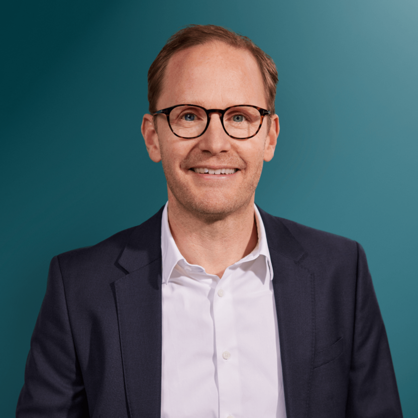Mann mit Brille und Anzug lächelt vor einem einfarbigen Hintergrund