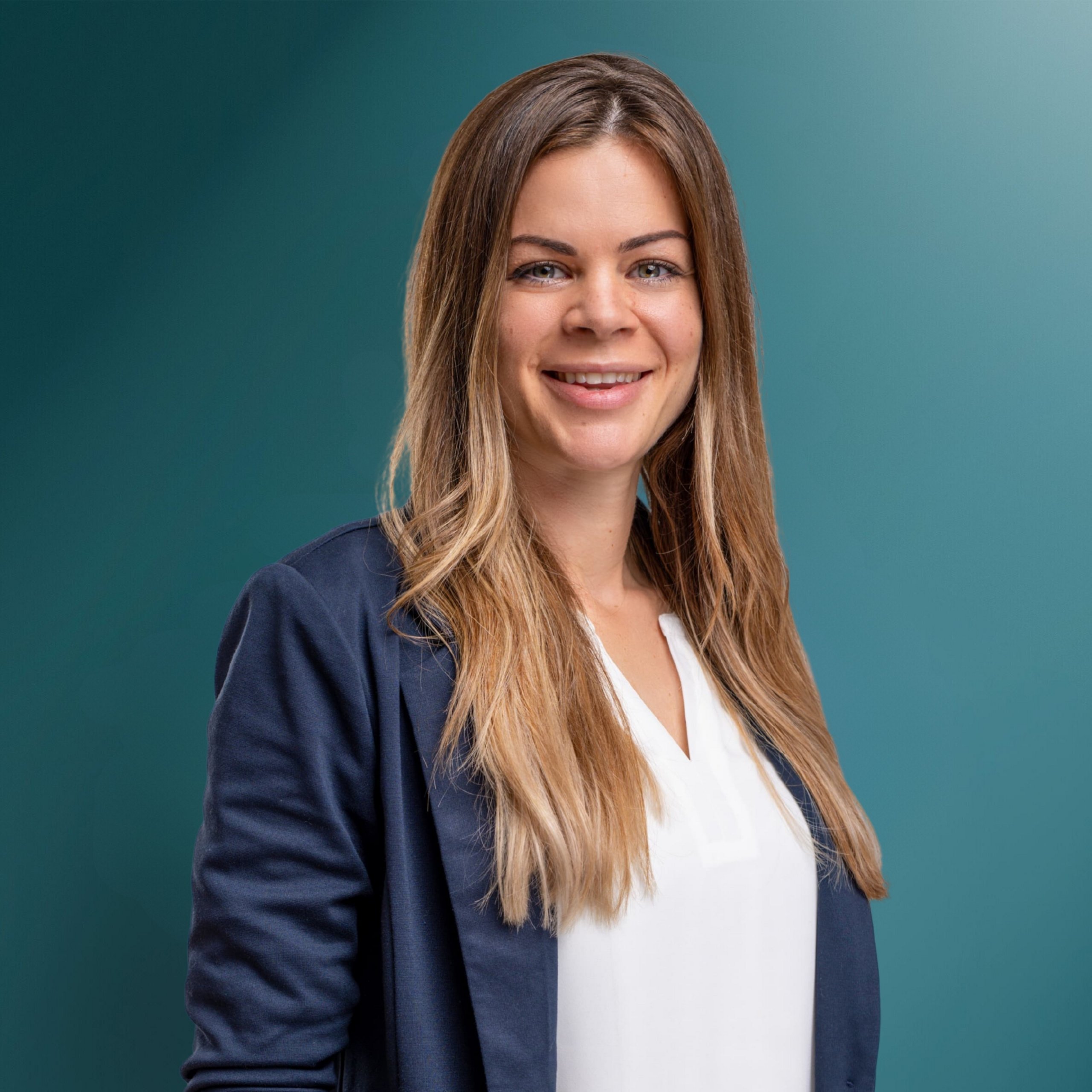 Professionelle Porträtaufnahme einer lächelnden Frau mit langen, hellen Haaren, die einen blauen Blazer und ein weißes Oberteil trägt, vor einem einfarbigen grünen Hintergrund