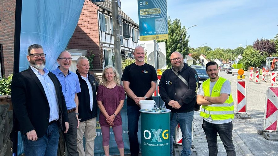 Gruppe von fünf Personen, die in Dortmund Wickede vor einem Banner der OXG steht, während im Hintergrund Bauarbeiten für Hochgeschwindigkeitsglasfaser stattfinden.