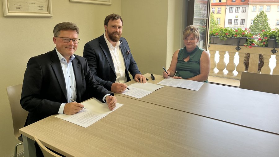 Drei Personen unterzeichnen an einem Tisch Dokumente zum Glasfaserausbau in der Region Weißenfels in einem hellen Raum mit Blick auf eine belebte Straße.