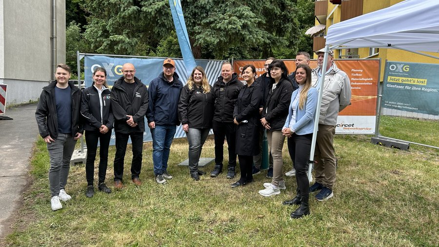 Eine Gruppe von zwölf Personen steht in der Stadt Suhl im Freien vor einem Banner, das Glasfaserprojekte bewirbt, umgeben von Bäumen und einer Wiese