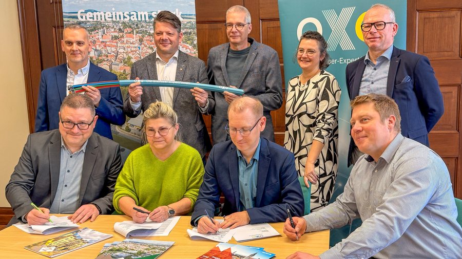 Mehrere Personen stehen und sitzen an einem Tisch, einige unterschreiben Dokumente, andere halten ein Glasfaserkabel; im Hintergrund OXG‑Banner und Stadtmotiv.