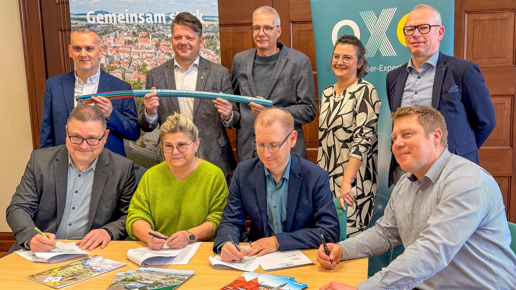 Mehrere Personen stehen und sitzen an einem Tisch, einige unterschreiben Dokumente, andere halten ein Glasfaserkabel; im Hintergrund OXG‑Banner und Stadtmotiv.