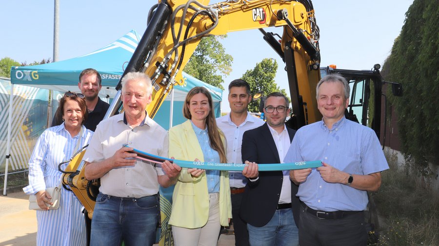 Eine Gruppe von sieben Personen draußen vor einem Bagger und einem Pavillion zum Gruppenfoto aufgereiht. Ein Teil der Gruppe hält gemeinsam ein Glasfaserkabel fest.