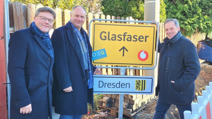 Drei Männer stehen lächelnd neben einem Verkehrsschild, das auf den Glasfaserausbau in Dresden hinweist