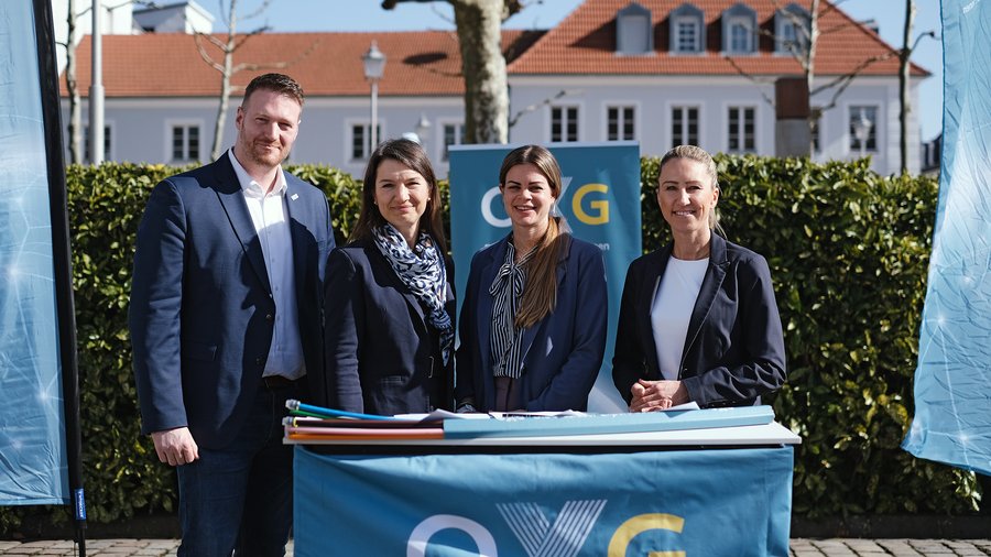 Vier Personen stehen draußen hinter einem Tisch mit OXG‑Logo, darauf Dokumente und ein petrolfarbenes Glasfaserkabel, im Hintergrund eine Hecke und Gebäude.