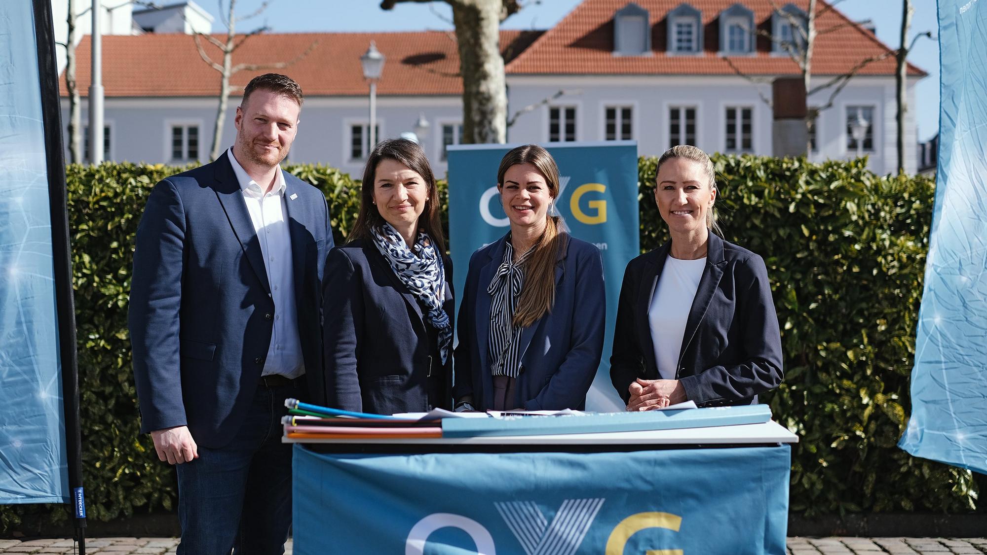 Vier Personen stehen draußen hinter einem Tisch mit OXG‑Logo, darauf Dokumente und ein petrolfarbenes Glasfaserkabel, im Hintergrund eine Hecke und Gebäude.