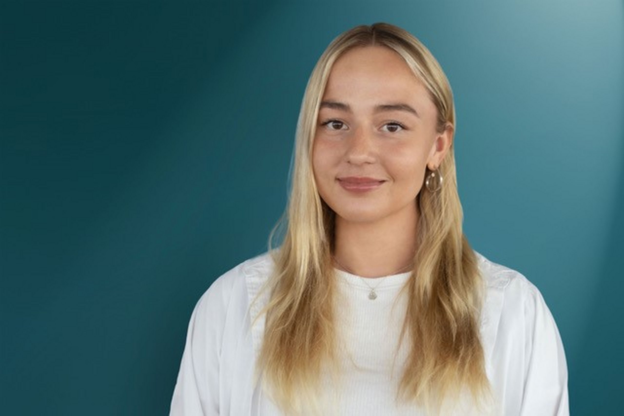 Porträt einer jungen Frau mit langen, blonden Haaren, die in einem hellen Oberteil vor einem türkisfarbenen Hintergrund steht