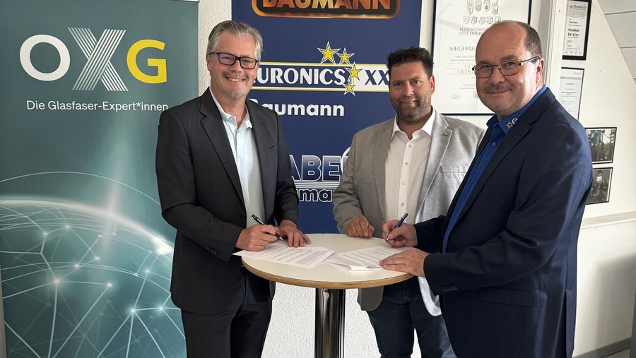 Drei Männer in Anzügen unterzeichnen Verträge an einem runden Tisch vor Firmenlogos und einem Banner der OXG, einem Anbieter für Glasfaserlösungen.
