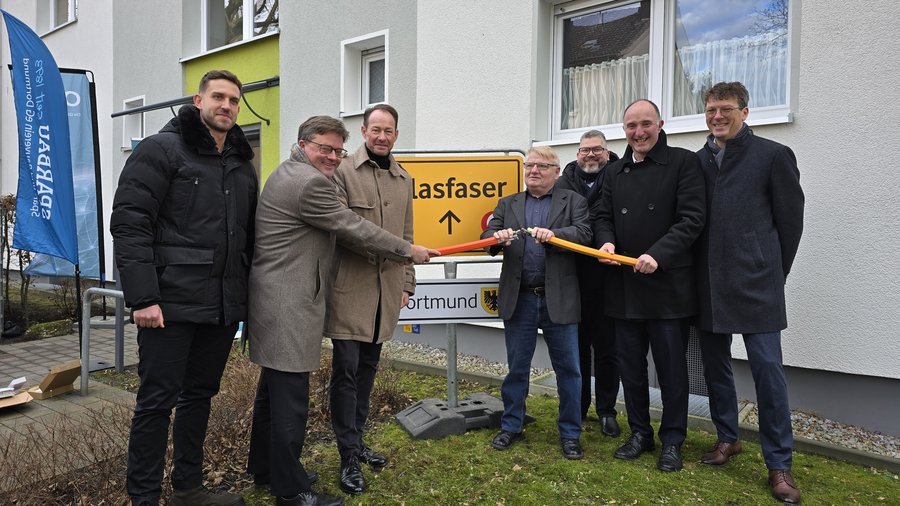 Personen stehen zusammen und halten gemeinsam ein orangefarbenes Kabel vor einem Schild, das auf die Verfügbarkeit von Glasfaser in Dortmund hinweist, während im Hintergrund ein Banner zu sehen ist.