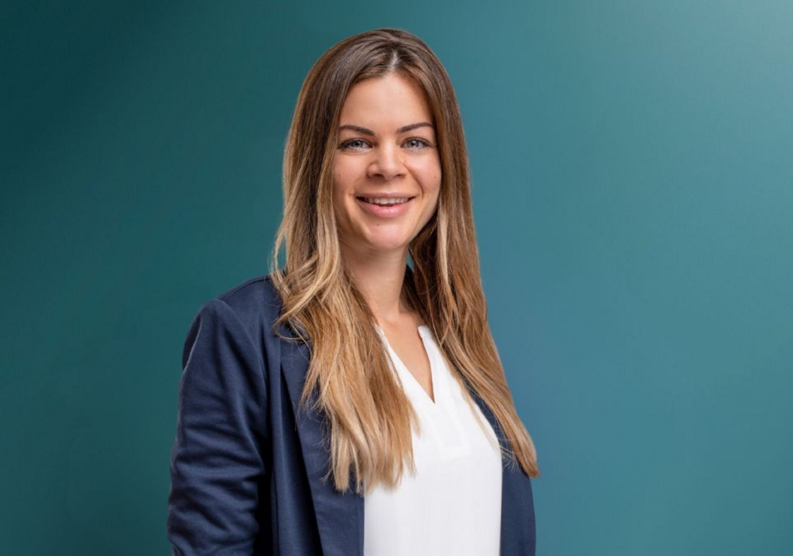 Porträt einer lächelnden Frau mit langen, glatten Haaren, die in einem Blazer und einem weißen Oberteil vor einem einfarbigen Hintergrund steht