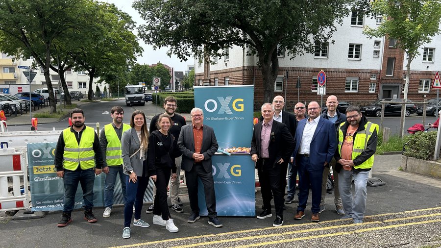 Die Gruppe von Fachleuten im Bereich Glasfasertechnik posiert vor einem Werbebanner der OXG und einer Baustelle in einer urbanen Umgebung in Koblenz.