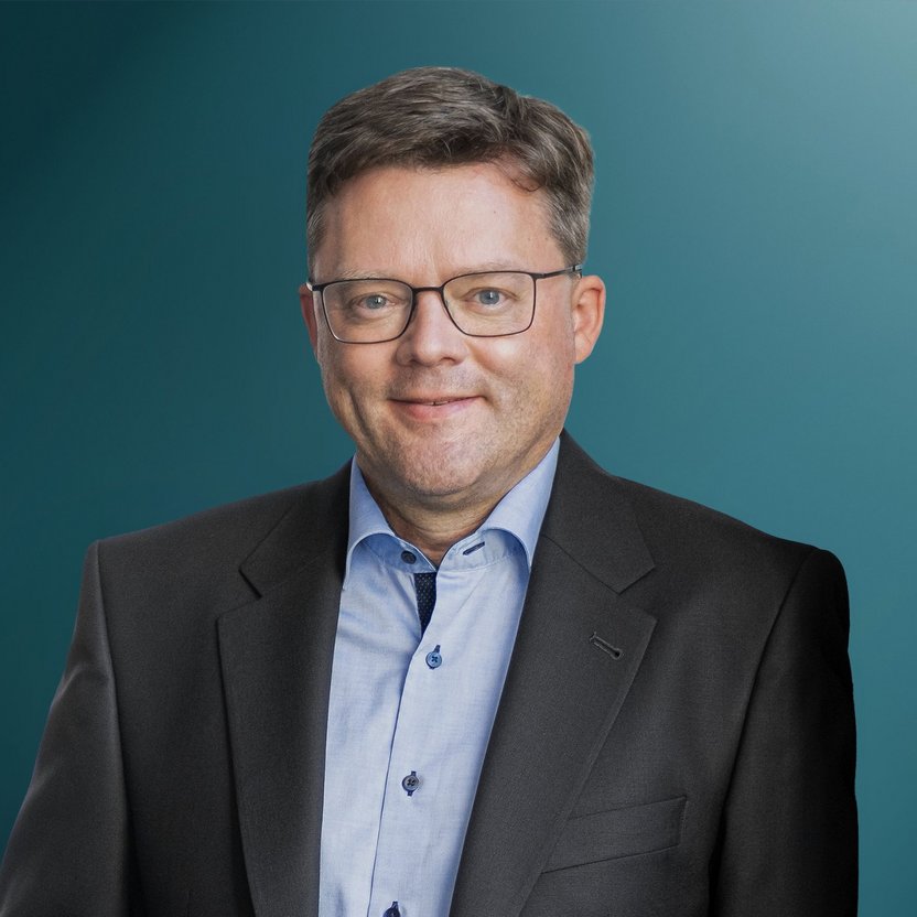 Ein Mann posiert in einem schwarzen Anzug mit einem hellblauen Hemd vor einem einfarbigen Hintergrund