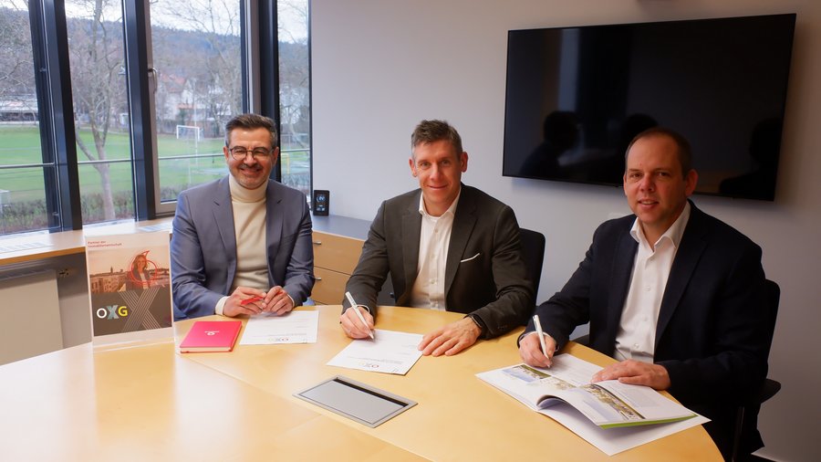 Drei Geschäftsleute sitzen an einem Tisch und unterschreiben Dokumente zur Partnerschaft mit dem Bauverein Breisgau, während im Hintergrund ein Fenster mit Blick auf eine grüne Wiese und einen Sportplatz sichtbar ist
